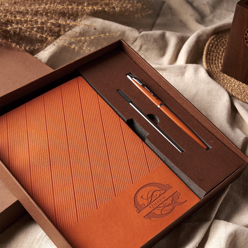 May include: A boxed gift set featuring a burnt orange leather-bound journal with vertical lines and the word "Queen" embossed on the lower right corner. A matching pen and refill are nestled in the box. The pen has a silver tip and a burnt orange barrel.
