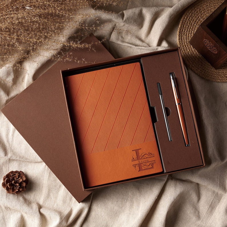 May include: A boxed gift set featuring a burnt orange leather-bound notebook with diagonal lines and a matching pen. The notebook has a personalized monogram. The set is presented in a brown box, ideal for gifting.