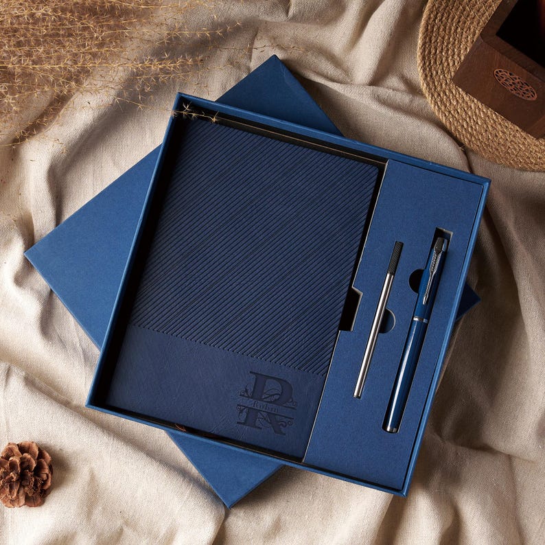May include: A navy blue gift set featuring a notebook and two pens. The notebook has a diagonal textured pattern and a monogram. The pens are silver and navy blue. The set is presented in a matching blue box.