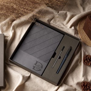 May include: A gift set in a dark gray box, featuring a notebook with diagonal lines and the name "DANIEL" in a decorative font, a pen, and a pen refill. The set is presented on a beige fabric surface, with dried plants and pine cones.