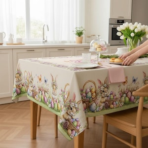 Op de afbeelding: Een rechthoekig paastafelkleed met een crèmekleurige achtergrond. De rand heeft illustraties van konijntjes, paaseieren, vlinders en bloemmotieven. Het tafelkleed ligt op een houten tafel in een goed verlichte keuken.
