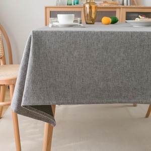 Puede incluir: Un mantel de lino gris cubre una mesa de madera. El mantel tiene un aspecto texturizado y cuelga de los bordes de la mesa. Una taza y un platillo blancos están sobre la mesa, junto con algunos objetos decorativos.