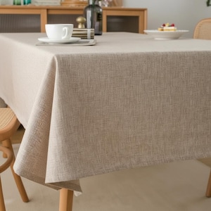 Könnte beinhalten: Ein beiges Leinentischtuch bedeckt einen Holztisch, auf dem eine weiße Tasse und Untertasse stehen. Das Tischtuch hat ein strukturiertes Aussehen und fällt über die Tischkanten. Ein kleiner Teller mit Essen ist im Hintergrund zu sehen.