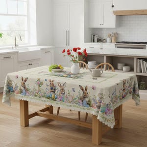 Op de afbeelding: Een rechthoekig paastafelkleed met een geschulpte rand. De crèmekleurige stof heeft een herhalend patroon van konijntjes, paaseieren en bloemmotieven in pastelkleuren. Het tafelkleed ligt op een houten tafel in een lichte keuken.