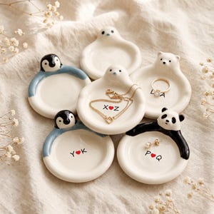 Könnte beinhalten: Fünf kleine, runde, cremefarbene Keramikschalen mit skulpturalen Tierfiguren wie Pinguinen, einem Panda und Eisbären. Die Schalen sind mit schwarzen Buchstaben und roten Herzsymbolen personalisiert und enthalten teilweise Schmuck wie einen Goldring.