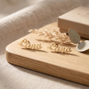 Puede incluir: Pendientes dorados con el nombre "Luna" en escritura cursiva, presentados sobre una superficie de madera. Los pendientes se muestran con flores secas y una hoja, con una caja de regalo en el fondo.