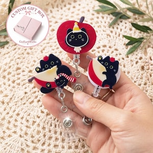 Op de afbeelding: Drie intrekbare badgehouders met zwarte katontwerpen. Eén toont een kat met een feesthoedje op een rode appel, een andere een kat met een rood-witte gestreepte sjaal, en de derde een kat die een rood-witte schijfje fruit vasthoudt.