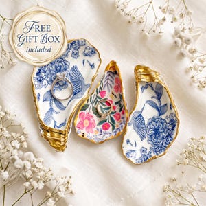 May include: Three decorative oyster shell dishes with gold-painted edges. Two shells feature blue and white floral and bird patterns, while the center shell displays a colorful floral design. Two silver rings rest inside the shells. Text reads FREE GIFT BOX included.