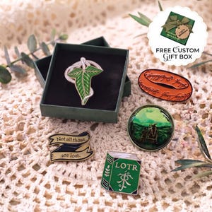 Puede incluir: Un conjunto de cinco pines de esmalte que incluye un broche de hoja verde en una caja oscura, un pin ovalado naranja con inscripciones, un pin circular de paisaje, un pin de cinta con el texto Not all those are lost y un pin de libro verde LOTR.