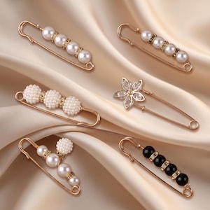 May include: A collection of six gold-tone safety pins featuring decorative accents. Two pins have four white pearls with crystal spacers, one has three textured pearl clusters, one has a crystal flower, one has mixed pearls, and one has four black beads.