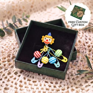 May include: A clown brooch featuring a yellow-haired clown head with a red nose, attached to multiple colourful safety pins. The pins are decorated with small striped spheres in red, white, yellow, and blue. The brooch is presented in a dark green box.