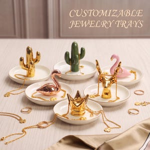 May include: A collection of white ceramic jewellery trays with gold rims, each featuring a central decorative figurine including a gold cactus, green cactus, pink flamingo, rose gold swan, gold deer head, and gold crown, with gold necklaces and rings.