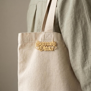 May include: A gold-colored pin with the names "Samuel & Olivia" in an elegant script font, with heart accents, is attached to a beige canvas tote bag. The bag has a simple design with cream-colored straps.