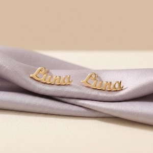 Puede incluir: Un par de pendientes dorados con el nombre "Luna" escrito en cursiva. Los pendientes se muestran sobre una tela suave de color lavanda.