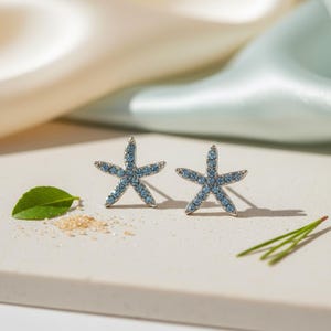 May include: A pair of silver starfish stud earrings, each adorned with light blue gemstones. The earrings are set against a neutral background with a green leaf and a scattering of sand, evoking a beach theme.
