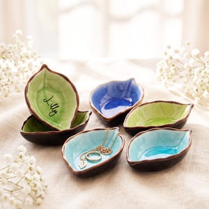 Puede incluir: Un conjunto de seis pequeños platos de cerámica con forma de hoja, con interiores brillantes en tonos verde claro, azul y turquesa con bordes de color marrón oscuro. Un plato verde está personalizado con el nombre Lilly en cursiva negra.