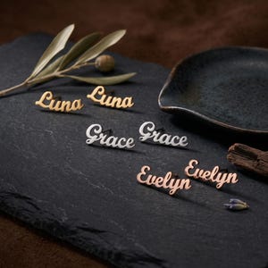 Può includere: Orecchini personalizzati con nomi come "Luna", "Grace" ed "Evelyn", in oro, argento e oro rosa. I nomi sono scritti in corsivo. Presentati su una superficie di ardesia scura con elementi decorativi.