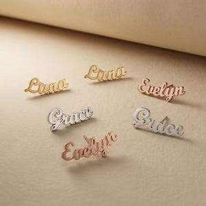 Puede incluir: Seis pendientes de botón con nombres en tonos oro, plata y oro rosa. Los nombres "Luna", "Grace" y "Evelyn" son visibles. Los pendientes están en una fuente cursiva y se muestran sobre un fondo neutro.