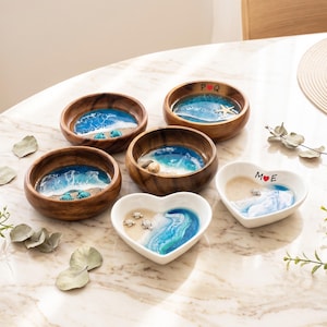 May include: A collection of four round wooden bowls and two heart-shaped white ceramic bowls featuring resin ocean art. The designs include sandy beaches, blue waves, and small sea creature figures. Two bowls have custom lettering with red heart icons.