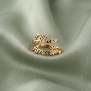 May include: A gold-colored ring with the names "Oliver" and "Amelia" in cursive script. The ring is set against a soft, sage green fabric background. The ring's design features two bands, one for each name.