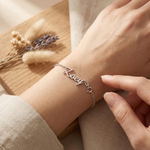 May include: A silver bracelet with the name "Zoey" in cursive script. The bracelet is delicate and features a small clasp. The image shows the bracelet on a wrist, with a neutral background.