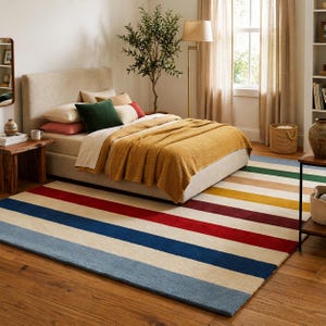 Puede incluir: Un dormitorio con una cama con cabecero beige, almohadas y una manta color mostaza. Una colorida alfombra a rayas en azul, rojo, beige, verde y amarillo cubre el suelo. También se ven una mesita de noche de madera y una lámpara de pie.