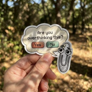 May include: A sticker featuring a speech bubble with the text "Are you overthinking this?" and a cartoon paperclip. The bubble has "Yes" and "No" buttons. The sticker is held in a hand against a blurred outdoor background.