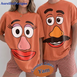 Pode incluir: Duas camisetas laranja com rostos de personagens de desenho animado. A camiseta da esquerda exibe um rosto com cílios longos, sombra roxa, nariz rosa e lábios vermelhos. A da direita mostra um rosto com um nariz laranja e um bigode preto.