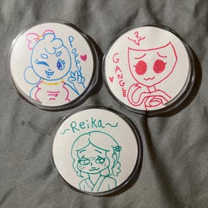 May include: Three round, clear-backed pins with hand-drawn cartoon characters. One pin features a blue character with the name "Poppy" and a pink heart. Another pin has a red character named "Gangle". The third pin has a green character named "Reika".