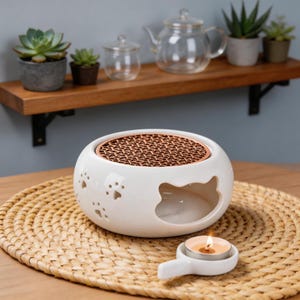 May include: A white ceramic cat-shaped oil warmer with a copper-colored top and paw print cutouts. A small tea light candle sits in a matching holder. The warmer is on a woven mat, with a shelf of plants and a glass teapot in the background.