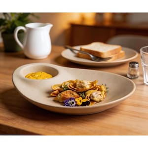 May include: A beige ceramic serving plate with a built-in sauce bowl, filled with yellow sauce, and a portion of ravioli garnished with flowers and herbs. Two slices of bread and a white pitcher are in the background.