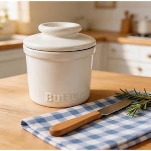 May include: A white ceramic butter dish with a lid, embossed with the word "Butter". A wooden-handled butter knife and a sprig of rosemary rest on a blue and white checkered cloth.
