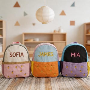 May include: Three colorful children's backpacks with embroidered names: SOFIA, JAMES, and MIA. Each backpack features a different color scheme, including pink, blue, and purple, with a pineapple pattern on the front pocket. The backpacks have a top handle and adjustable straps.
