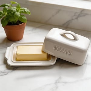 Może przedstawiać: Biała ceramiczna maselniczka z pokrywką i uchwytem, obok prostokątnego naczynia z kostką masła. Na przodzie pokrywki wytłoczony napis "BUTTER". Mała roślina doniczkowa w tle.