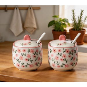 Op de afbeelding: Twee bijpassende keramische suikerpotten met deksels en lepels. De kommen zijn wit met een roze bloemenpatroon en roze accenten op de deksels. De lepels zijn wit. De kommen staan op een houten oppervlak.
