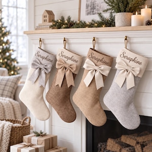 Könnte beinhalten: Vier Weihnachtsstrümpfe hängen in einer Reihe, jeder mit dem Namen Seraphine in dunkler Schreibschrift bestickt. Die Strümpfe bestehen aus strukturiertem Stoff in Creme, Hellbraun, Beige und Grau und sind jeweils mit einer passenden Stoffschleife verziert.