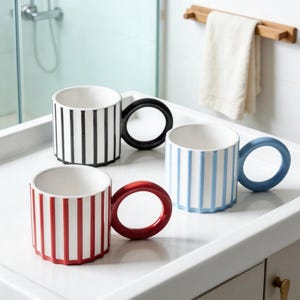 Puede incluir: Tres tazas de cerámica con patrones de rayas verticales en negro, rojo y azul, cada una con un asa circular del mismo color. Las tazas son blancas por dentro y están sobre una superficie blanca. Artículos de cocina decorativos.