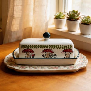 Może przedstawiać: Biała ceramiczna maselniczka z pokrywką i ciemnozielonym guzikiem. Naczynie zdobią ilustracje czerwonych grzybów i zielone akcenty liści. Całość stoi na talerzu w kwiatowy wzór, z ciemnozieloną obwódką.