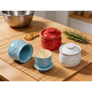 May include: A collection of ceramic butter dishes in various colors, including red, white, and blue. Each dish has a lid and the word "Butter" embossed on the side. One blue dish is open, filled with butter. The dishes sit on a wooden surface.