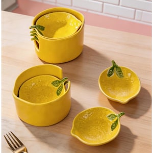 Puede incluir: Un juego de platos de cerámica amarillos con forma de limón. El juego incluye dos recipientes más grandes con insertos en forma de limón y dos platos pequeños con forma de limón. Cada limón tiene una hoja verde y un detalle de tallo.