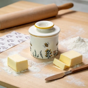Pode incluir: Uma manteigueira de cerâmica branca com borda amarela e ilustrações de abelhas e flores. O prato está ao lado de um rolo de madeira, manteiga e uma faca de manteiga em uma tábua de corte de madeira.