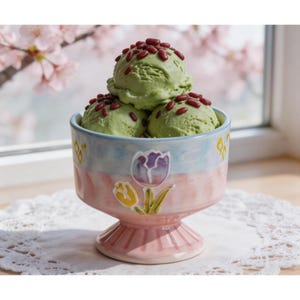 May include: A decorative ice cream bowl filled with three scoops of green ice cream, topped with red sprinkles. The bowl is pink with a blue and yellow floral design. The bowl is sitting on a white doily.