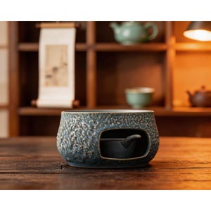 Puede incluir: Un calentador de té de cerámica azul con un exterior texturizado y una pequeña taza de té de color oscuro en el interior. El calentador se encuentra sobre una superficie de madera, con una estantería de artículos relacionados con el té en el fondo.