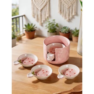 May include: A pink ceramic serving set with a strawberry design. The set includes a holder, small bowls, and spoons. The bowls are white with pink interiors and red dot accents. The spoons have a white handle. The set is on a wooden table.