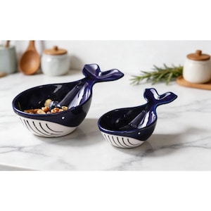 May include: Two ceramic whale-shaped bowls. The bowls are navy blue with white bellies and a tail handle. The larger bowl contains nuts and berries. The bowls are on a white marble surface.