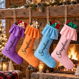 Könnte beinhalten: Vier weiche Weihnachtsstrümpfe in Lila, Orange, Hellblau und Rosa, jeweils mit gerüschtem Umschlag, Jute-Aufhängeschlaufe und dem Namen Seraphine in schwarzer Schreibschrift. Jeder Strumpf enthält ein kleines Geschenk mit einer Schleife.