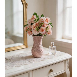 Puede incluir: Un jarrón rosa con diseño de corazón contiene un ramo de flores color melocotón con hojas verdes. El jarrón está sobre una mesa blanca con un camino de mesa de encaje, junto a un frasco de perfume y un espejo con marco dorado.