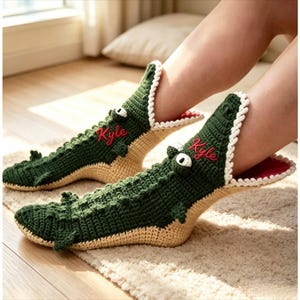 Peut inclure: Une paire de chaussons en crochet vert foncé en forme de crocodiles avec un dos texturé, une bordure de dents blanches, une doublure intérieure rouge et le prénom Kyle brodé en écriture rouge sur le côté de chaque chausson.