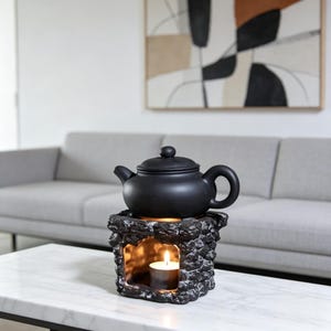 Puede incluir: Una tetera negra se encuentra sobre un calentador decorativo con forma de piedra y una vela encendida en su interior. La tetera tiene un cuerpo redondeado, un pico curvo y un asa en forma de bucle. El calentador tiene un acabado texturizado gris y negro.