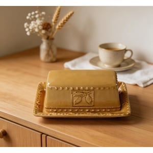 May include: A mustard-yellow ceramic butter dish with a lid, featuring a rectangular shape and a decorative embossed design of leaves. The dish sits on a matching rectangular base. A cup of coffee and a small vase with dried flowers are in the background.
